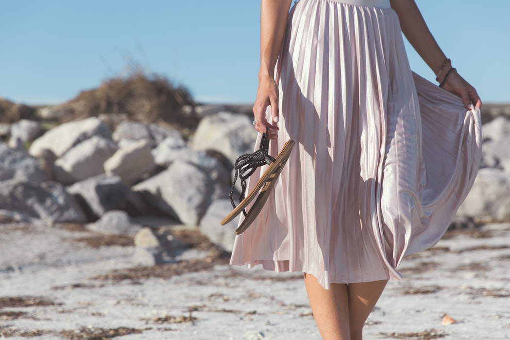 Kobieta idąca po plaży, w ręku trzyma sandały i torbę - ubrana jest w plisowaną spódnicę do kolan
