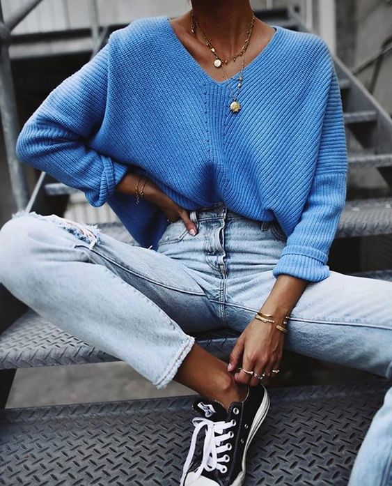 Kobieta ubrana w błękitny sweter oversize, podarte jeansy oraz trapki